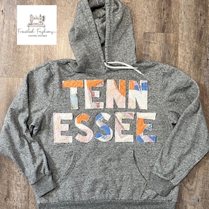 Op de afbeelding: Grijze hoodie met het woord "TENNESSEE" in kleurrijke patchworkstof op de borst. De hoodie heeft een capuchon met trekkoord en een voorzak. Het merklogo staat in de linkerbovenhoek.