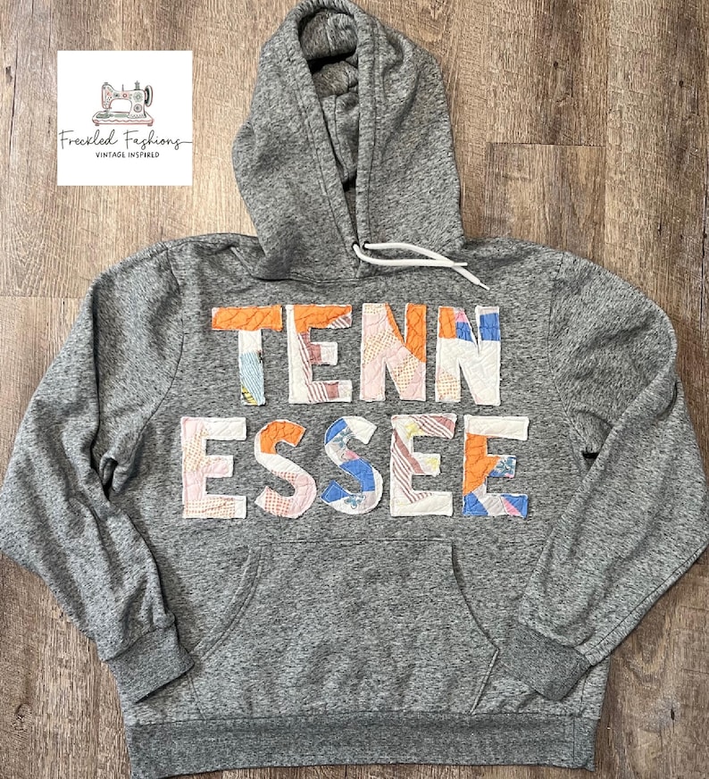 Op de afbeelding: Grijze hoodie met het woord "TENNESSEE" in kleurrijke patchworkstof op de borst. De hoodie heeft een capuchon met trekkoord en een voorzak. Het merklogo staat in de linkerbovenhoek.