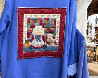 Puede incluir: Una sudadera azul bígaro de cuello redondo con una gran aplicación de tela acolchada cuadrada en el pecho. La aplicación representa una muñeca con un gorro blanco y un lazo azul, sentada junto a un gato sobre un fondo de patchwork colorido.