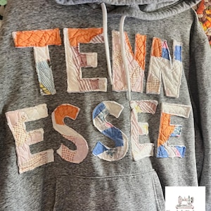 Op de afbeelding: Grijze hoodie met het woord "TENNESSEE" op de borst. De letters zijn gemaakt van patchworkstof in verschillende kleuren, waaronder oranje, roze, blauw en wit. De hoodie heeft een capuchon met trekkoord en een voorzak.