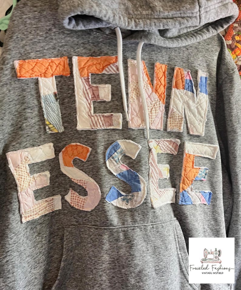 Op de afbeelding: Grijze hoodie met het woord "TENNESSEE" op de borst. De letters zijn gemaakt van patchworkstof in verschillende kleuren, waaronder oranje, roze, blauw en wit. De hoodie heeft een capuchon met trekkoord en een voorzak.