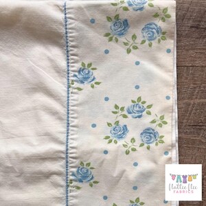 Op de afbeelding: Witte stof met een blauw bloemenpatroon en blauwe stippen. De stof heeft een blauwe gestikte rand. De stof is gelabeld met "Nattie Nic Fabrics".