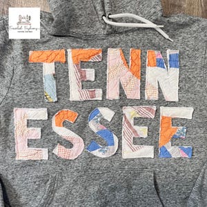 Op de afbeelding: Grijze hoodie met het woord "TENNESSEE" in kleurrijke patchworkstof op de borst. De hoodie heeft een capuchon met trekkoord en een voorzak. In de linkerbovenhoek staat een logo met de tekst "Freckled Fashions Vintage Inspired".