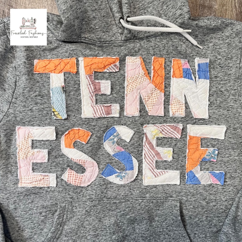 Op de afbeelding: Grijze hoodie met het woord "TENNESSEE" in kleurrijke patchworkstof op de borst. De hoodie heeft een capuchon met trekkoord en een voorzak. In de linkerbovenhoek staat een logo met de tekst "Freckled Fashions Vintage Inspired".