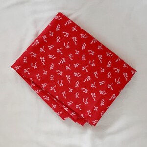 May include: Folded red bandana with a repeating white bow pattern. The fabric is a vibrant red, and the bows are evenly spaced across the surface. The bandana is neatly folded, showcasing the pattern and color.