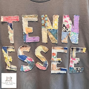 Op de afbeelding: Donkergrijze sweatshirt met het woord "TENNESSEE" in patchworkstof. Elke letter is gemaakt van verschillende stoffen met patronen in rood, blauw, geel en bloemmotieven. Een klein logo met een naaimachine staat linksonder.
