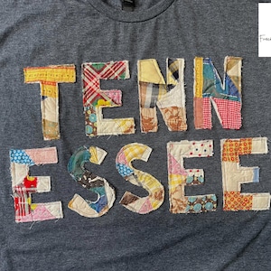 Op de afbeelding: Een grijs t-shirt met het woord "TENNESSEE" op de voorkant. De letters zijn gemaakt van verschillende stoffen met patronen in tinten geel, rood, blauw en bruin. Het shirt heeft een ronde hals en korte mouwen.