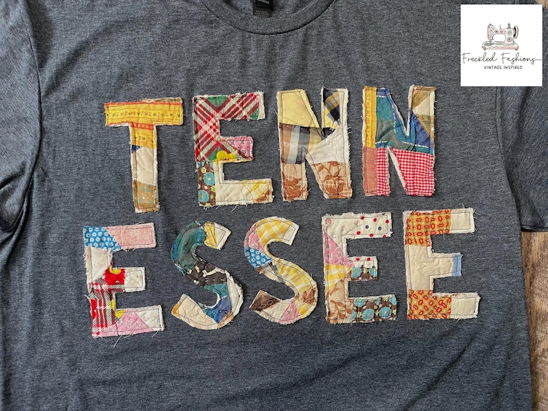 Op de afbeelding: Een grijs t-shirt met het woord "TENNESSEE" op de voorkant. De letters zijn gemaakt van verschillende stoffen met patronen in tinten geel, rood, blauw en bruin. Het shirt heeft een ronde hals en korte mouwen.
