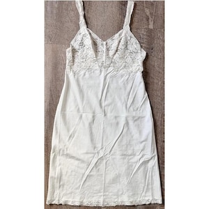 Könnte beinhalten: Ein weißes Unterkleid mit V-Ausschnitt und Spaghettiträgern. Das Oberteil hat einen zarten Spitzeneinsatz, und der Saum ist mit passender Spitze verziert. Das Kleid ist aus einem weichen, leichten Stoff gefertigt.