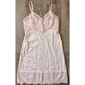 Könnte beinhalten: Ein hellrosa Unterkleid mit verstellbaren Trägern. Das Oberteil hat einen zarten Spitzeneinsatz, und der Saum ist mit passender Spitze verziert. Das Unterkleid ist aus einem weichen, fließenden Stoff gefertigt.