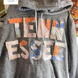 Op de afbeelding: Grijze hoodie met de tekst "TENN ESSE" in patchworkstof. De letters zijn in verschillende kleuren en patronen, waaronder oranje, roze, blauw en wit. De hoodie heeft een capuchon met trekkoord en een voorzak.