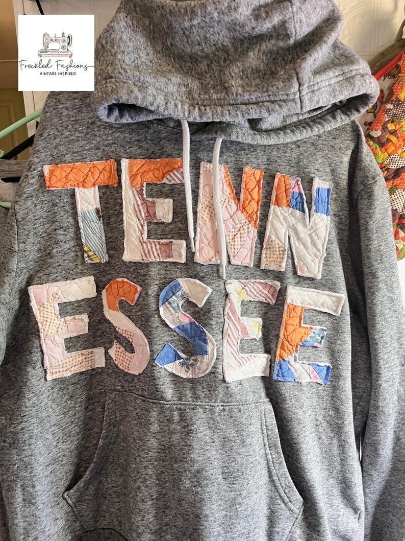 Op de afbeelding: Grijze hoodie met de tekst "TENN ESSE" in patchworkstof. De letters zijn in verschillende kleuren en patronen, waaronder oranje, roze, blauw en wit. De hoodie heeft een capuchon met trekkoord en een voorzak.