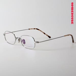 以下が含まれることがあります： シルバーフレームのメガネ、楕円形のレンズ。テンプルは茶色とタン色のべっ甲模様です。メガネは白い背景に置かれています。「404REDCUBE」の文字が右上に赤色で表示されています。
