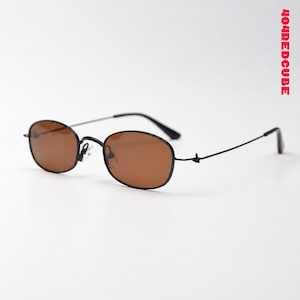 May include: Oval sunglasses with thin black metal frames and brown tinted lenses. The temples are slender and black, featuring a small knot detail near the hinges. The text 404REDCUBE is printed vertically in red on the right side of the frame.