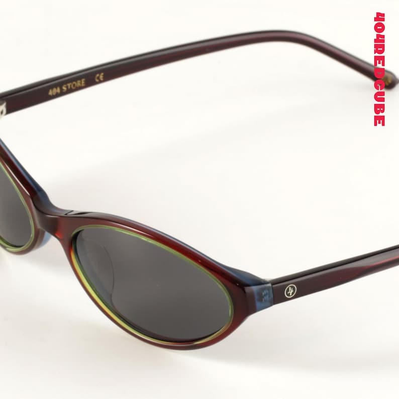 May include: Oval-shaped sunglasses with dark lenses and a burgundy frame. The frame has a thin green and blue accent around the lenses. The text "404 STORE CE" is printed on the left arm. The right arm features a logo.