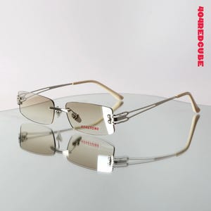 Puede incluir: Gafas sin montura con un tinte marrón claro. Las monturas son plateadas con patillas beige. El texto "404STORE" es visible en la lente. El texto "404REDCUBE" está impreso verticalmente en el lado derecho.