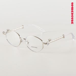 Könnte beinhalten: Silberfarbene Brille mit ovalen Gläsern. Die Fassungen haben dekorative Akzente an den Bügeln und Scharnieren, mit kleinen, funkelnden Verzierungen. Der Text "404STORE" ist auf dem Glas sichtbar. Der Markenname "404REDCUBE" ist rot gedruckt.