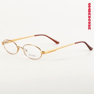 Puede incluir: Gafas ovaladas doradas con patillas marrones. Las gafas tienen una montura delicada y lentes transparentes. El texto "404STORE" está impreso en la lente. La imagen está sobre un fondo blanco.