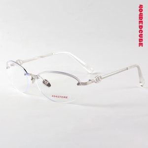 Puede incluir: Gafas sin montura con lentes ovaladas y montura de metal plateado. Las patillas tienen un diseño decorativo cerca de las bisagras. El texto "404STORE" está impreso en la lente inferior derecha.