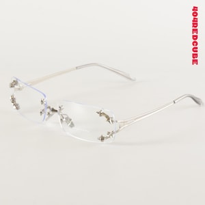 Puede incluir: Gafas sin montura de color plateado con lentes transparentes. Las gafas presentan adornos decorativos en forma de estrella en la montura y las patillas. El nombre de la marca es visible en la lente. El diseño es moderno y elegante.