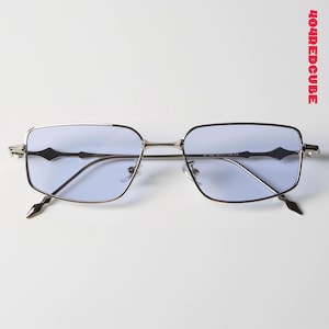 Puede incluir: Gafas de sol con montura plateada y lentes azul claro. Las monturas rectangulares tienen una forma de diamante negro decorativa en las patillas y puntas puntiagudas. El texto "404REDCUBE" está impreso en rojo en el lado derecho.