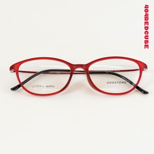Op de afbeelding: Rode, ovale bril met zwarte pootjes. De monturen hebben de tekst "ULTEM by sable" op de linker lens en "404STORE" op de rechter lens. De tekst "404REDCUBE" is verticaal aan de rechterkant gedrukt.