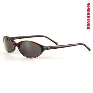 May include: Oval-shaped sunglasses with dark lenses and a brown frame. The frame has a green and blue accent around the lenses and thin brown arms. The brand name "404REDCUBE" is printed vertically in red on the right side.