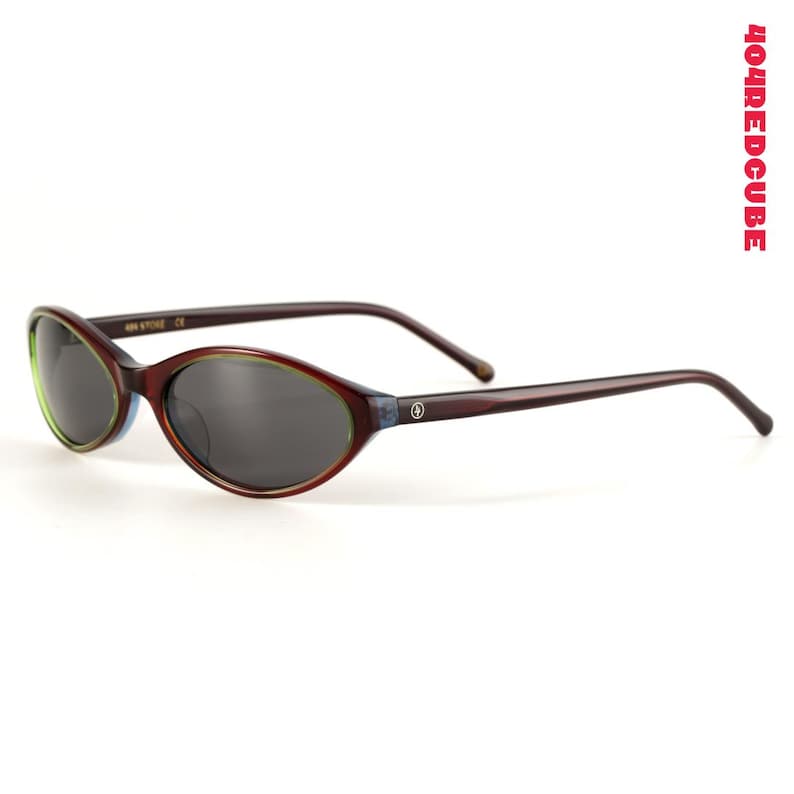 May include: Oval-shaped sunglasses with dark lenses and a brown frame. The frame has a green and blue accent around the lenses and thin brown arms. The brand name "404REDCUBE" is printed vertically in red on the right side.