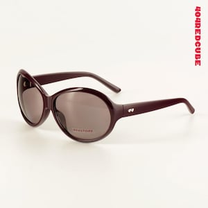 Może przedstawiać: Owalne okulary przeciwsłoneczne w głębokim bordowym odcieniu. Soczewki są w stonowanym brązowym kolorze, a oprawka posiada mały dekoracyjny detal na zausznikach. Tekst "404STORE" jest wydrukowany na prawej soczewce. Tekst "404REDCUBE" jest wydrukowany pionowo po prawej stronie.