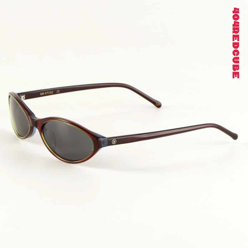 May include: Oval-shaped sunglasses with dark lenses and a dark brown frame. The frame has a subtle blue and green accent around the lenses. The temples are long and slender. The text "404 STORE CE" is printed on the left temple.