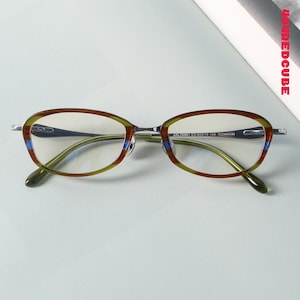 Può includere: Occhiali ovali con montatura fantasia marrone, verde e blu. Gli occhiali hanno ponte e aste argentate e lenti trasparenti. Il testo "ARL78881 C3 62019 148 TITANIUM" è stampato sulla montatura.