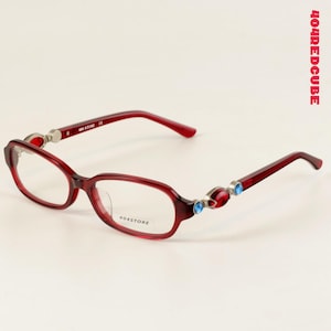 Puede incluir: Gafas rojas con montura ovalada. Las patillas presentan detalles plateados y adornos de joyas rojas y azules. El texto "404STORE" es visible en la lente.