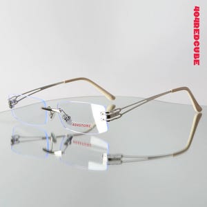 Puede incluir: Gafas sin montura con monturas de metal plateado y patillas beige. Las lentes rectangulares tienen el texto "404STORE" en la lente inferior derecha. Las gafas se reflejan en una superficie brillante.