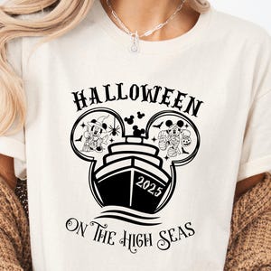 May include: Cream-colored t-shirt with a black graphic featuring a ship with Mickey Mouse ears, the text "HALLOWEEN" above, and "ON THE HIGH SEAS" below. The ship has the year "2025" on it. Mickey Mouse is dressed in Halloween costumes.
