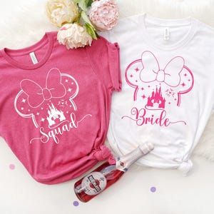 Puede incluir: Dos camisetas, una rosa y otra blanca, con un diseño inspirado en Minnie Mouse. La camiseta rosa dice "Squad" y la camiseta blanca dice "Bride". Una botella de vino espumoso está en primer plano.