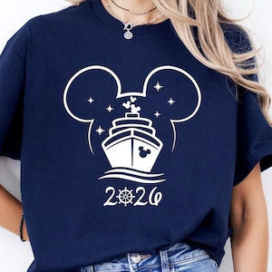 May include: Navy blue t-shirt featuring a white graphic of a cruise ship inside a Mickey Mouse head outline, with stars and the year 2026. The design includes a steering wheel symbol. The shirt is a casual wear item.