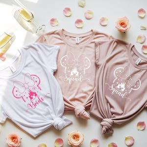 May include: Three t-shirts with a Minnie Mouse-inspired design. One white shirt has "Bride" in pink script, while the other two are pink and feature "Squad". The shirts are knotted at the bottom and surrounded by rose petals and champagne glasses.