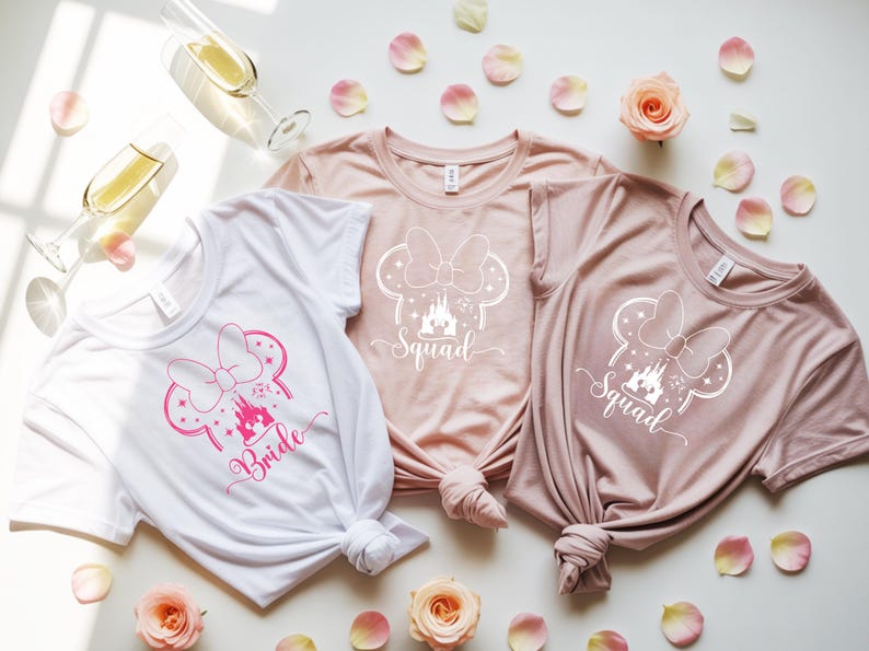 May include: Three t-shirts with a Minnie Mouse-inspired design. One white shirt has "Bride" in pink script, while the other two are pink and feature "Squad". The shirts are knotted at the bottom and surrounded by rose petals and champagne glasses.