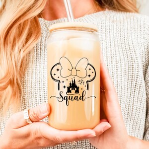 May include: A clear glass tumbler with a wooden lid and a straw, filled with a light-colored beverage. The tumbler features a black graphic of a bow, mouse ears, a castle, stars, and the word "Squad".
