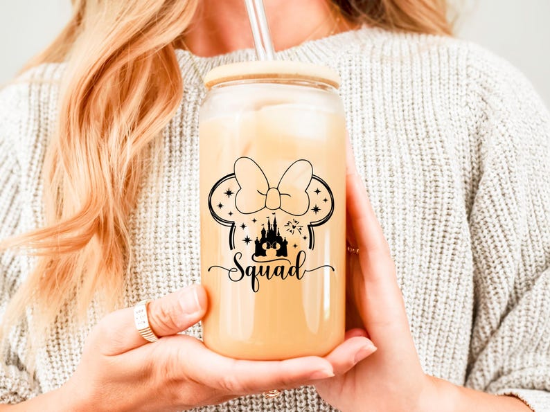 May include: A clear glass tumbler with a wooden lid and a straw, filled with a light-colored beverage. The tumbler features a black graphic of a bow, mouse ears, a castle, stars, and the word "Squad".