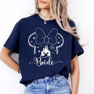May include: Navy blue t-shirt featuring a white graphic of a Minnie Mouse bow, ears, and a castle, with the word "Bride" in a script font. The shirt is made of a soft material and is suitable for a wedding.
