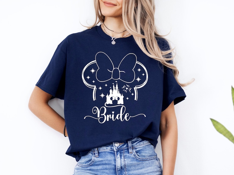 May include: Navy blue t-shirt featuring a white graphic of a Minnie Mouse bow, ears, and a castle, with the word "Bride" in a script font. The shirt is made of a soft material and is suitable for a wedding.