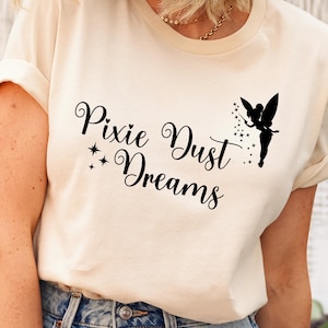 May include: A white t-shirt with the text "Pixie Dust Dreams" in black cursive lettering. A silhouette of a fairy with a wand is to the right of the text.