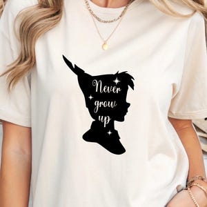 Może przedstawiać: Biała koszulka z czarną sylwetką Piotrusia Pana i tekstem "Never grow up".