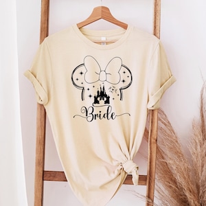 May include: A light beige t-shirt with a black graphic of Minnie Mouse ears, a bow, a castle, and the word "Bride" in a script font. The shirt is hanging on a wooden ladder.