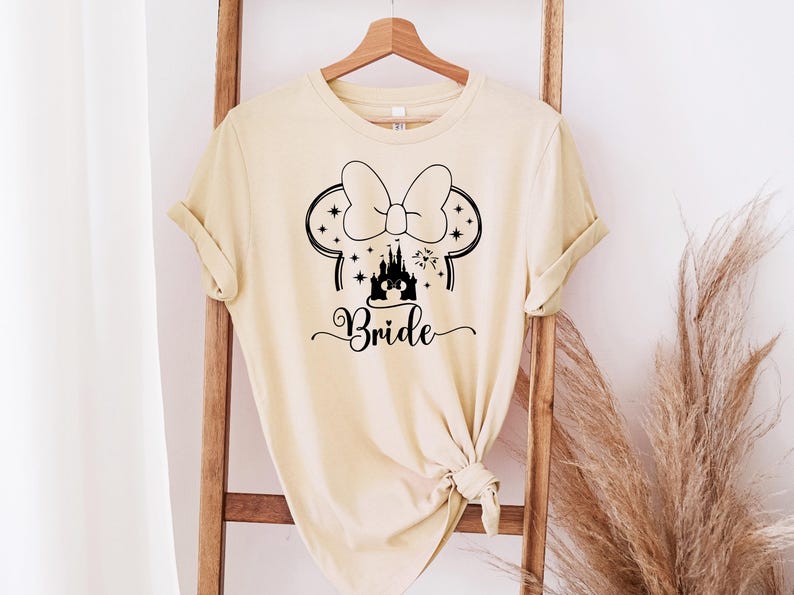 May include: A light beige t-shirt with a black graphic of Minnie Mouse ears, a bow, a castle, and the word "Bride" in a script font. The shirt is hanging on a wooden ladder.