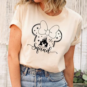 May include: A cream-colored t-shirt featuring a black graphic of a bow, mouse ears, a castle, and the word "Squad" in a script font. The shirt is worn with blue jeans.
