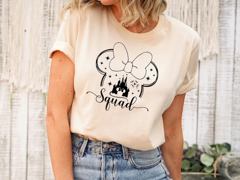 May include: A cream-colored t-shirt featuring a black graphic of a bow, mouse ears, a castle, and the word "Squad" in a script font. The shirt is worn with blue jeans.