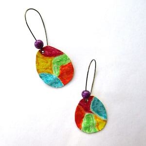 May include: A pair of abstract art earrings. Each earring features a colorful, oval-shaped pendant with a mix of red, yellow, green, and blue hues. A small, purple bead is attached to the pendant, and a thin, dark wire forms the earring hook.