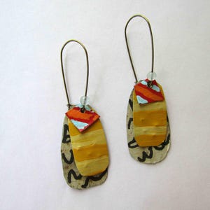 May include: Artisan earrings with a distinctive, artistic design. Each earring has a layered construction with a painted yellow and orange oval shape, accented by a small, colourful diamond shape. The earrings hang from bronze-coloured ear wires and include a small bead.